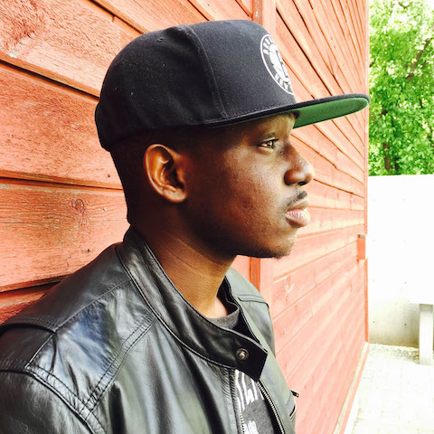 A profile shot of Adekunle wearing a jacket and baseball cap standing outside.