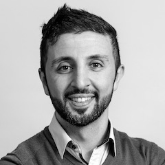 A black and white portrait of Amir who has a neatly trimmed beard and a Tintin-like haircut wearing a jumper over a shirt unbuttoned at the neck.
