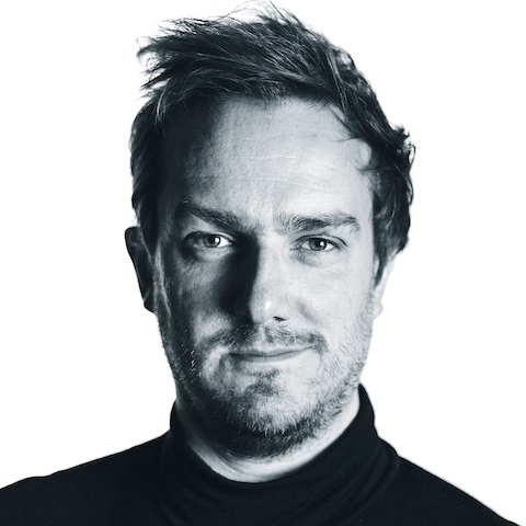 A monochrome portrait of John with a wry smile on his face, wearing a black turtleneck in the clichéd design tradition.