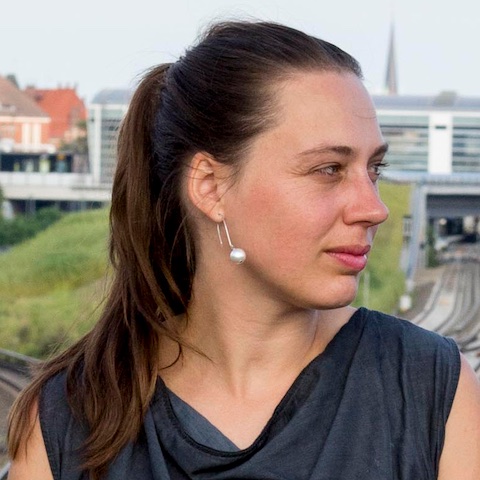 Julia looking off to the side as she stands on an overpass above train tracks in an urban setting.