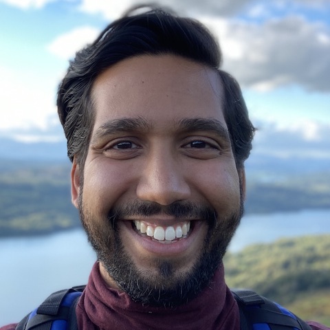 The beaming bearded face of Videha standing in front of the beautiful landscape of a riverbank.