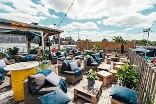 An outdoor rooftop bar with wooden tables and comfy cushions on the seats.